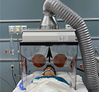 person lying on a bed with a ventilator hood surrounding them. A plexiglass case holds up a silver hood with a large foil vent draped to the side.